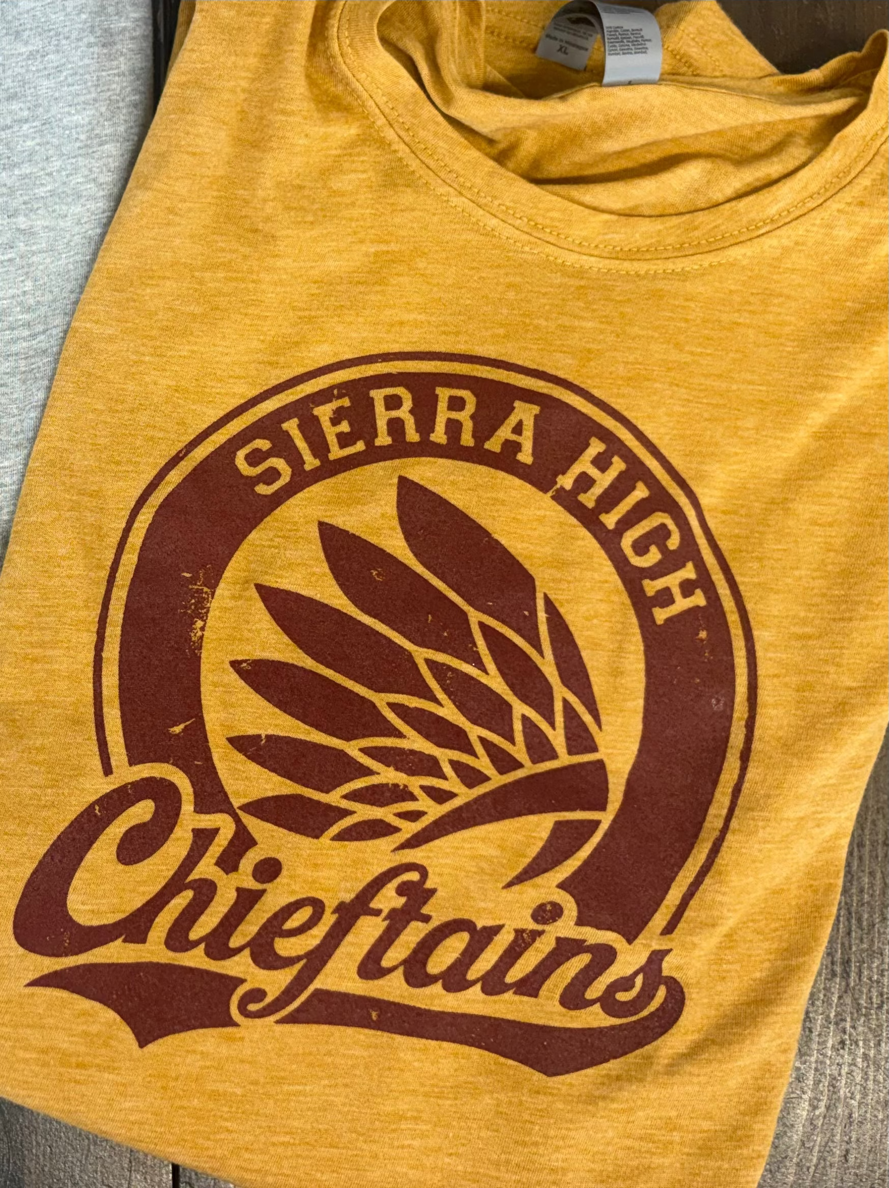 A close-up of a heather mustard yellow t-shirt featuring a distressed dark brown graphic print of a wheat stalk inside a circular emblem with the text 'Chiefs' and 'SIERRA HIGH'.