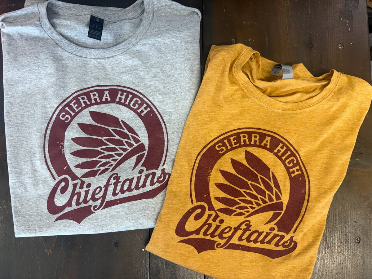 Two folded graphic t-shirts displayed on a dark wooden surface. One is light heather gray and the other is mustard yellow, both featuring a distressed maroon circular 'SIERRA HIGH Chieftains' graphic with a feather and eagle design.